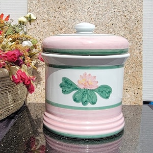 May include: A white ceramic canister with a pink lid and green trim. The canister has a painted design of a pink flower with green leaves.