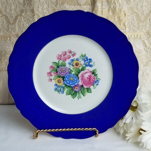 May include: A blue and white porcelain dinner plate with a gold rim. The center of the plate features a floral design with pink, blue, yellow, and purple flowers.