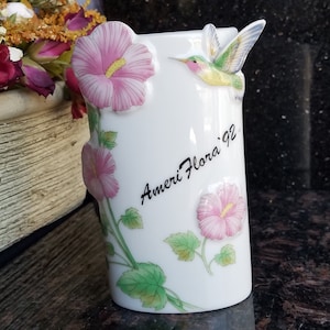 Puede incluir: Jarrón de cerámica blanca con un colibrí y flores de hibisco rosas. El jarrón tiene el texto "Ameri Flora '92" escrito en él.