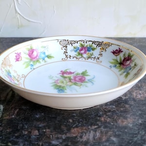 May include: A white porcelain bowl with a floral design. The bowl features pink and purple roses, blue flowers, and gold trim. The rim has a decorative gold pattern. A classic piece for serving.