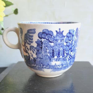 May include: A white teacup with a blue Willow pattern design. The cup features a handle and a detailed landscape scene with pagodas, trees, and a fence. The cup is sitting on a dark surface.
