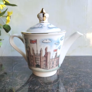 May include: A white ceramic teapot with a lid, featuring a detailed illustration of a castle with British flags against a blue sky. The teapot has a gold-colored finial and trim, and a curved handle and spout.