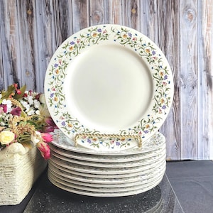Puede incluir: Una pila de 10 platos de cena de cerámica blanca con un diseño floral en tonos azules, amarillos y verdes. Los platos tienen un borde dorado.