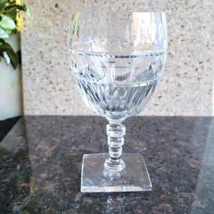 May include: A clear crystal wine glass with etched detailing. The glass features a square base, a stem with a stacked bead design, and a bowl with horizontal etched bands. The glass is designed for serving wine.