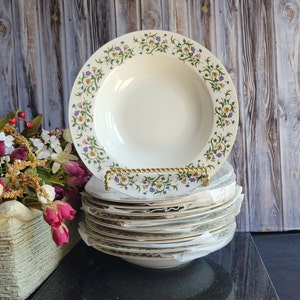 Puede incluir: Una pila de cuencos de sopa de cerámica blanca con un patrón floral. El cuenco superior tiene un borde dorado y un diseño floral con hojas verdes, flores moradas, amarillas y azules.