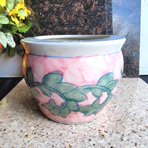 May include: A round ceramic planter featuring a white rim and a body decorated with a soft pink watercolor-style wash. A hand-painted green vine pattern with detailed leaves wraps horizontally around the center of the planter's exterior surface.