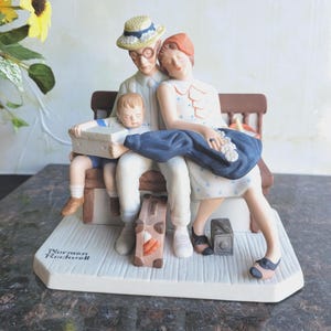 May include: A ceramic figurine depicting a family of three seated on a bench. The father wears a straw hat and glasses, the mother has red hair, and the child holds a box. The base is white with the text "Norman Rockwell".