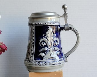 Op de afbeelding: Een blauwe en witte keramische bierkan met een tinnen deksel en handvat. De kan heeft een bloemmotief en is versierd met een wit en blauw patroon.