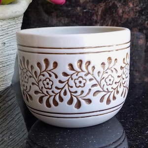 May include: A white ceramic planter featuring a repeating embossed floral vine pattern in a metallic bronze finish. The design includes horizontal bronze bands encircling the top and bottom edges of the cylindrical vessel, creating a clean look.