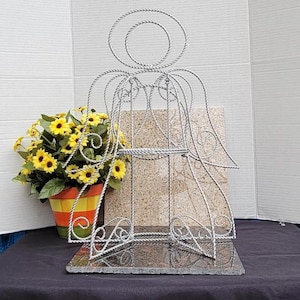May include: A silver wire angel-shaped plant stand with a granite base. The stand has two shelves and is decorated with a swirl design. A potted plant with yellow flowers is in front of the stand.