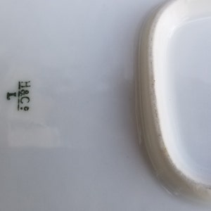 Puede incluir: Bandeja rectangular blanca para servir con un borde elevado. La bandeja tiene una marca verde en la parte inferior que dice "H&C.  L."