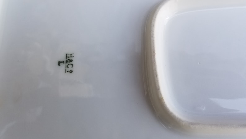 Puede incluir: Bandeja rectangular blanca para servir con un borde elevado. La bandeja tiene una marca verde en la parte inferior que dice "H&C.  L."