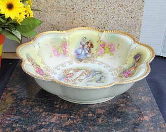 Könnte beinhalten: Eine große, weiße Porzellanschale mit einem gewellten Rand und einem goldenen Rand. Die Schale ist mit einem Blumenmuster und einer Szene von zwei Personen in einem Garten verziert. Die Schale steht auf einer dunkelbraunen Oberfläche.
