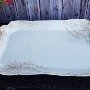 May include: A white porcelain serving tray with a gold rim and floral design. The tray has a scalloped edge and two handles.