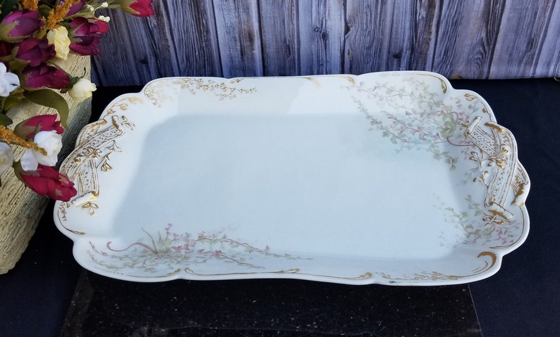Puede incluir: Una bandeja de servir de porcelana blanca con un borde dorado y un dise&ntilde;o floral. La bandeja tiene un borde festoneado y dos asas.