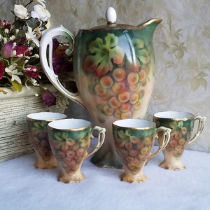 May include: A vintage porcelain tea set with a grape motif. The set includes a pitcher and four cups with handles. The pitcher has a lid and a gold trim. The cups have a gold rim and a delicate design.