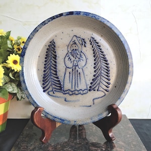 May include: A stoneware plate with a blue and white design. The plate features a central image of a robed figure between two stylized trees. The rim has a blue border. The plate is displayed on a wooden stand.