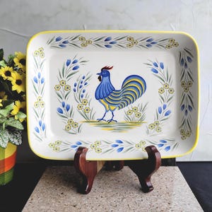 May include: Rectangular ceramic serving dish with a hand-painted rooster design. The rooster is blue and yellow, surrounded by floral accents. The dish has a yellow border and is displayed on a wooden stand.
