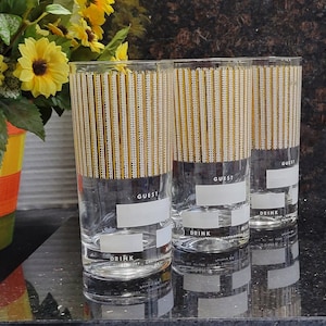 May include: Three clear glass drinking glasses with a gold and white striped pattern. The glasses have the word "GUEST" and "DRINK" printed on them.