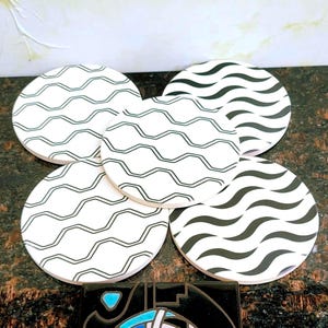 May include: A set of five circular coasters featuring a white background with black wavy line patterns. Three coasters display thin, outlined wave designs, while two coasters feature thicker, solid black wave patterns arranged in horizontal rows.