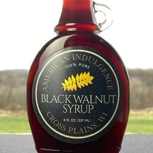 May include: A glass bottle of Black Walnut Syrup from American Indulgence. The dark syrup is visible through the clear glass. The label is black with gold lettering and a gold leaf design. The bottle contains 8 fl oz (237 ml).