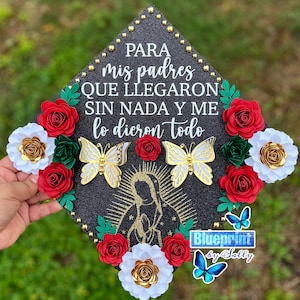 May include: A black graduation cap decorated with red and white paper flowers, gold butterflies, and a gold silhouette of the Virgin Mary. The cap has the text "PARA mis padres QUE LLEGARON SIN NADA Y ME lo dieron todo" and "Blueprint by Sally".