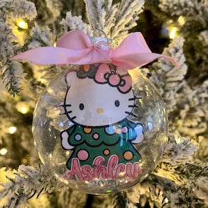Pode incluir: Enfeite de Natal de vidro transparente com um design Hello Kitty, um laço rosa e o nome "Ashley" em escrita rosa. O enfeite é decorado com um vestido verde com bolinhas rosa e laranja e o ano de 2024.