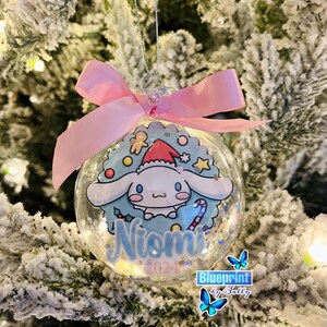 Pode incluir: Enfeite de Natal transparente com um design de personagem de desenho animado, um laço rosa e fio prateado. O enfeite apresenta um personagem azul e branco usando um chapéu de Papai Noel, com o nome "Niomi" e o ano "2024" em azul. O enfeite é preenchido com glitter.