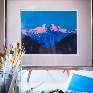 Op de afbeelding: Een ingelijste foto van een besneeuwde bergketen met een roze en blauwe lucht. De foto staat op een houten ezel met een palet en kwasten op de voorgrond.
