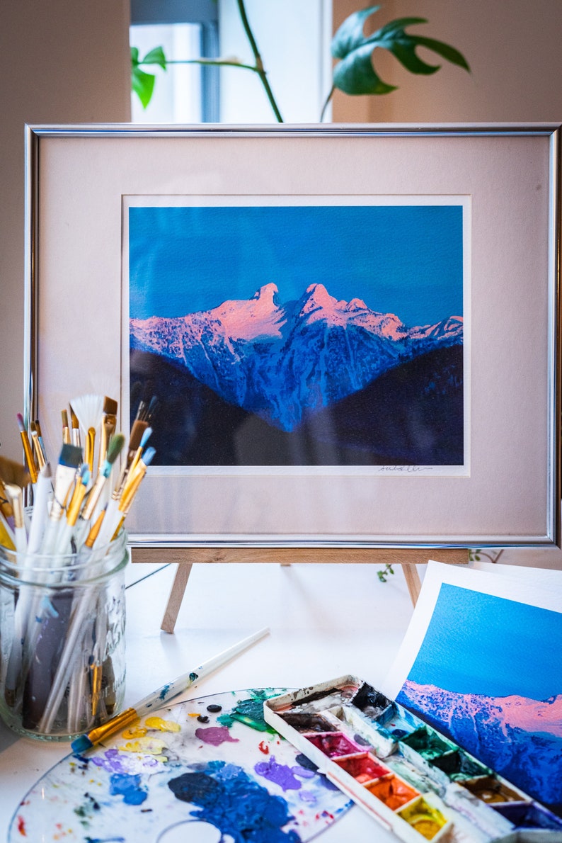 Op de afbeelding: Een ingelijste foto van een besneeuwde bergketen met een roze en blauwe lucht. De foto staat op een houten ezel met een palet en kwasten op de voorgrond.