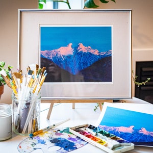 Op de afbeelding: Een ingelijste print van een bergketen met een roze en blauwe lucht. De print staat op een houten ezel en op de voorgrond staan ​​penseel, een palet en een pot verf.