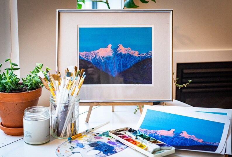 Op de afbeelding: Een ingelijste print van een bergketen met een roze en blauwe lucht. De print staat op een houten ezel en op de voorgrond staan ​​penseel, een palet en een pot verf.