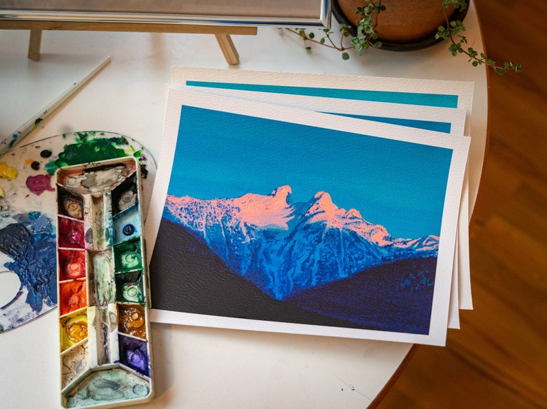 Op de afbeelding: Een print van een bergketen met een roze en blauwe kleurstelling. De bergen zijn bedekt met sneeuw en de lucht is een levendig blauw.