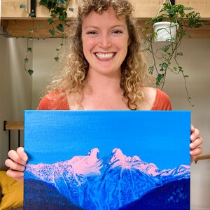 Op de afbeelding: Een schilderij van een bergketen met besneeuwde toppen tegen een helderblauwe lucht. De bergen zijn geschilderd in tinten blauw en paars, met roze accenten op de sneeuw. Het schilderij is op canvas en wordt vastgehouden door een persoon.