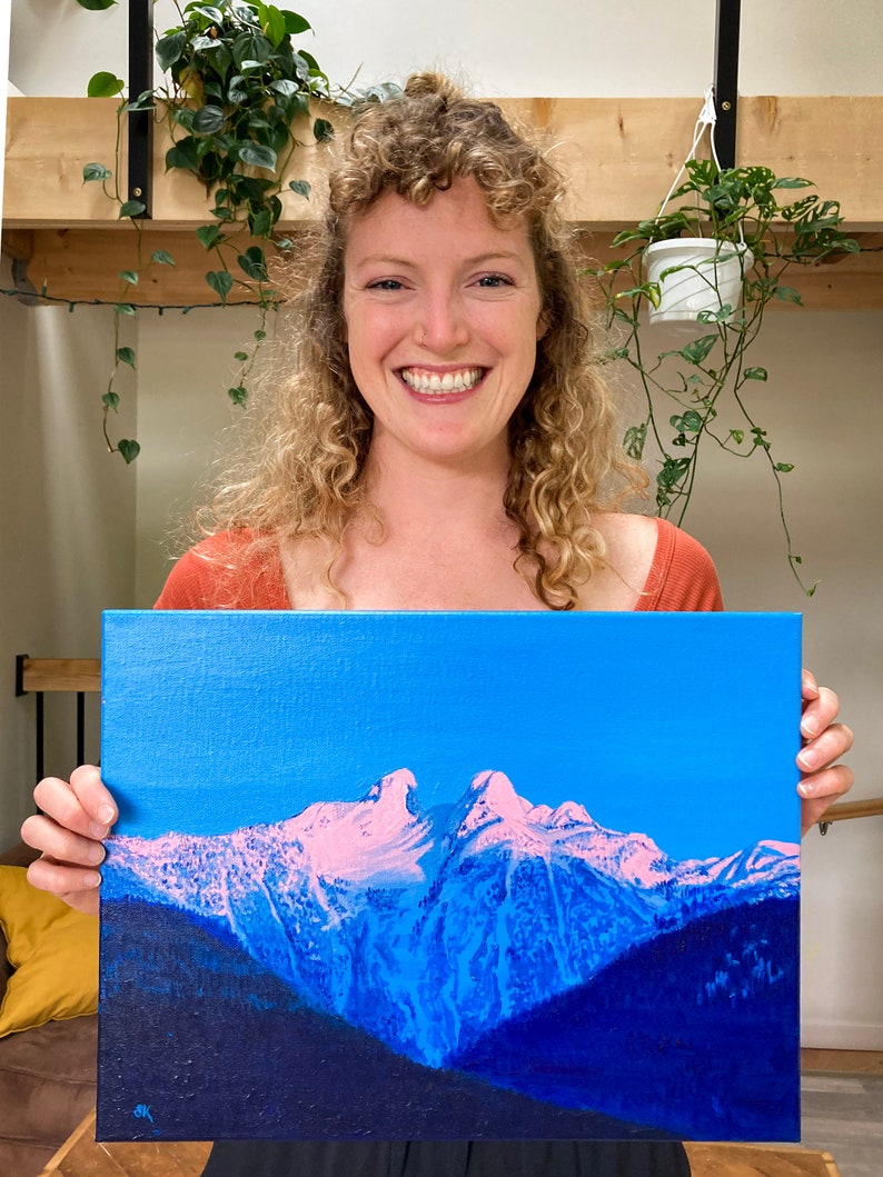 Op de afbeelding: Een schilderij van een bergketen met besneeuwde toppen tegen een helderblauwe lucht. De bergen zijn geschilderd in tinten blauw en paars, met roze accenten op de sneeuw. Het schilderij is op canvas en wordt vastgehouden door een persoon.