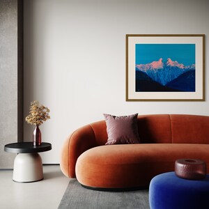 Op de afbeelding: Een modern woonkamerinterieur met een oranje fluwelen bank, een blauwe fluwelen poef en een ingelijste foto van een bergketen met een roze en blauwe lucht.