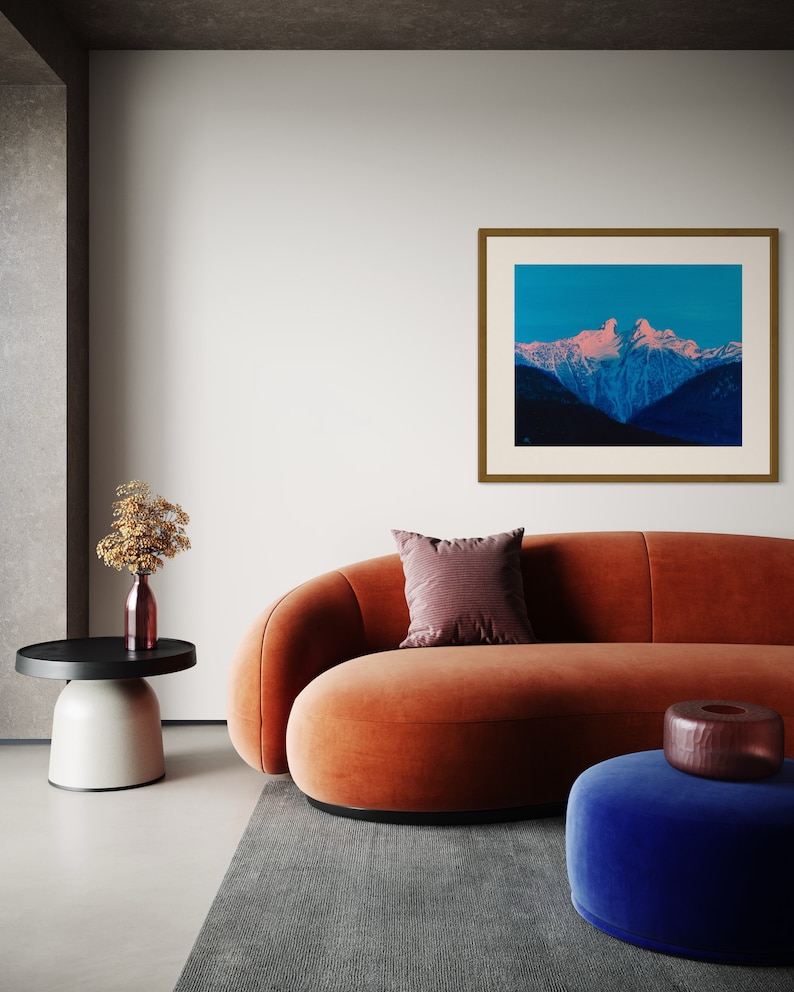Op de afbeelding: Een modern woonkamerinterieur met een oranje fluwelen bank, een blauwe fluwelen poef en een ingelijste foto van een bergketen met een roze en blauwe lucht.
