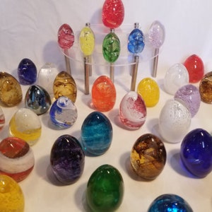 May include: A collection of colorful glass eggs with various patterns and textures. The eggs are arranged in a circular pattern on a white surface.