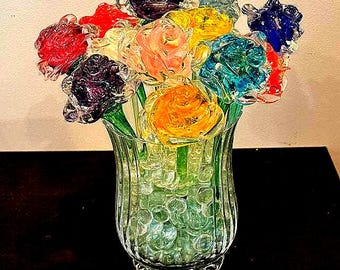 May include: A glass vase filled with colorful glass roses. The roses are in shades of red, pink, yellow, blue, and purple. The vase is clear glass with a textured surface.