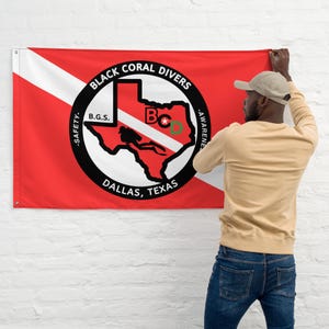 Puede incluir: Una bandera roja y blanca con un círculo negro que contiene el texto "Black Coral Divers", "B.G.S.", "Dallas, Texas", y un contorno del estado de Texas con un buceador en su interior. El texto "Safety" y "Awareness" está a cada lado del círculo. La bandera está siendo colgada en una pared de ladrillo blanco por una persona.