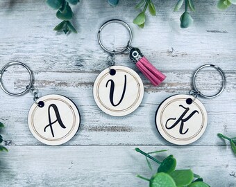 Puede incluir: Tres llaveros de madera con las letras A, V y K. Cada llavero tiene un anillo de llave plateado y un pequeño borla.