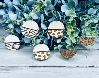 May include: Six pairs of wooden earrings with a white background and a brown animal print design. The earrings are hanging from a leafy green plant.