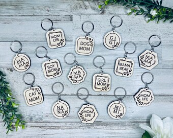 Op de afbeelding: Een verzameling van 14 houten sleutelhangers met verschillende ontwerpen en tekst. De sleutelhangers bevatten een verscheidenheid aan zinnen, waaronder "Mom Life", "Boy Mom", "Girl Mom", "Mama Bear", "Cat Mom", "Nana", "Mi Mi" en "Dog Mom". De sleutelhangers zijn allemaal gemaakt van natuurlijk hout en hebben een rustieke, handgemaakte uitstraling.
