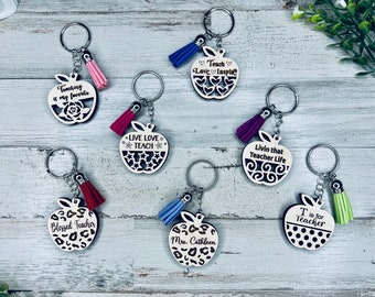 Op de afbeelding: Acht houten sleutelhangers in de vorm van een appel met verschillende ontwerpen en spreuken. Elke sleutelhanger heeft een klein kwastje. De spreuken zijn: "Onderwijs is mijn favoriet", "Leer Liefde Inspireer", "Leef Liefde Leer", "Leef dat lerarenleven", "Gezegende leraar", "Mevrouw Cathleen", "T is voor leraar".