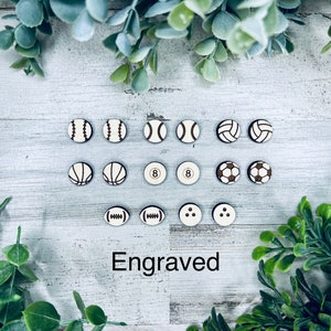 Pode incluir: Uma cole&ccedil;&atilde;o de 12 ornamentos redondos de madeira com v&aacute;rios designs esportivos, incluindo beisebol, basquete, v&ocirc;lei, futebol e uma bola de bilhar. Os ornamentos s&atilde;o brancos com detalhes marrons e a palavra "Gravado" est&aacute; escrita abaixo.