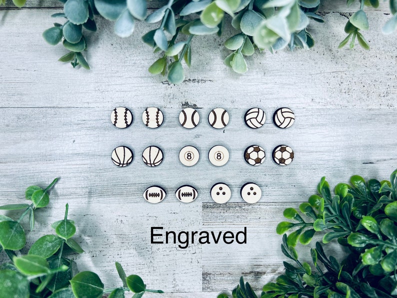 Pode incluir: Uma cole&ccedil;&atilde;o de 12 ornamentos redondos de madeira com v&aacute;rios designs esportivos, incluindo beisebol, basquete, v&ocirc;lei, futebol e uma bola de bilhar. Os ornamentos s&atilde;o brancos com detalhes marrons e a palavra "Gravado" est&aacute; escrita abaixo.