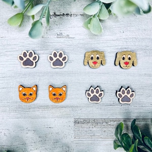 Pode incluir: Seis brincos de madeira com diferentes desenhos de animais e pegadas. Os brincos são pintados em laranja, marrom e cinza. As pegadas são cinzas e os animais são um gato e um cão.