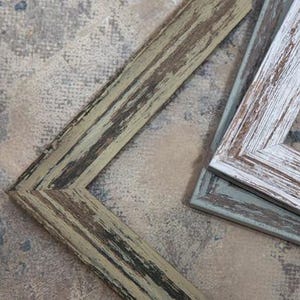 May include: Close-up of several wooden picture frames with distressed finishes. The frames are in various colours, including a weathered green, white, and black. The frames are angled, showcasing their textured surfaces and rustic aesthetic.