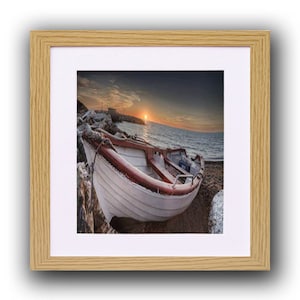 May include: A framed print showing a small white boat with red detailing, beached on a rocky shore. The image depicts a sunset over the sea, with the sun low on the horizon. The frame is a light wood colour.