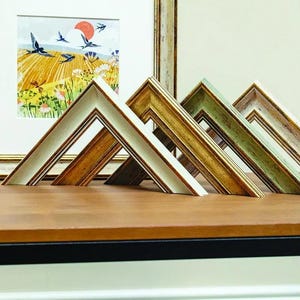 May include: Four L-shaped picture frame corner samples displayed in a row. The samples feature varied finishes including off-white, metallic gold, muted sage green, and a weathered wood tone, each with intricate moulding details and inner gold trim.